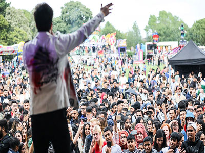 A photograph of a man singing from a stage. There is a large crowd in front of him and fairground rides are in the background.. Link to Do Good, Feel Good - Middlesbrough Mela set up,  Friday 15 August 2025.