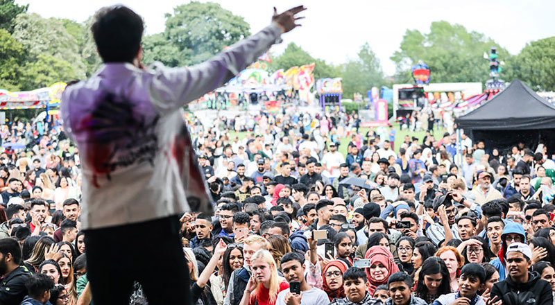 A photograph of a man singing from a stage. There is a large crowd in front of him and fairground rides are in the background.. Link to A photograph of a man singing from a stage. There is a large crowd in front of him and fairground rides are in the background..