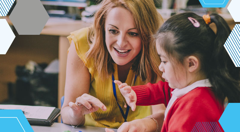 A teacher and a primary student in a red jumper interacting at a desk in abrightly lit classroom