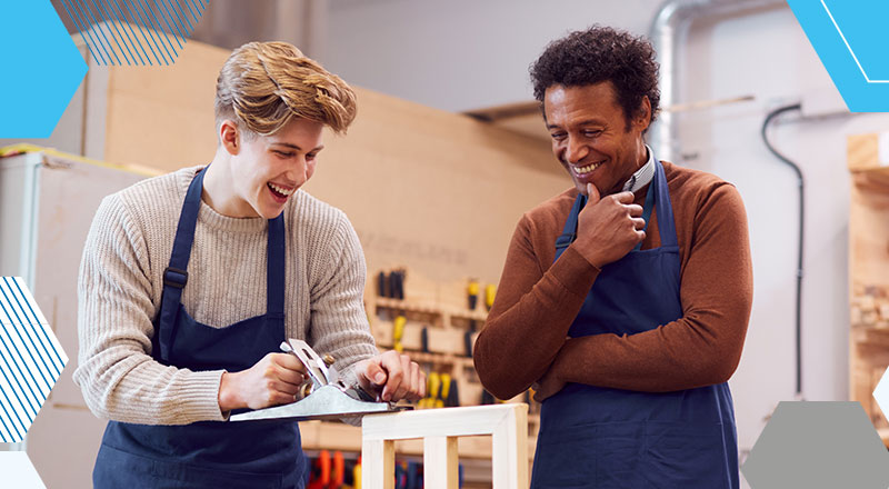 A teenage student is woodworking while a teacher oversee his work. Both are laughing